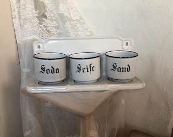 Op de afbeelding: Een wit geëmailleerd wandrek met drie cilindrische bakjes met blauwe randen. Elk bakje is voorzien van zwarte gotische letters met de woorden Soda, Seife en Sand, horizontaal gerangschikt op een rechthoekige basis.