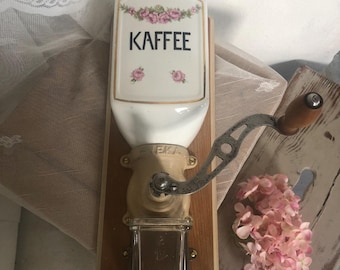 Op de afbeelding: Een wandkoffiemolen met een witte keramische trechter met roze bloemaccenten en het woord KAFFEE in zwarte letters. Het apparaat is voorzien van een metalen zwengel met een houten knop, een beige metalen maalmechanisme en een glazen opvangbak.