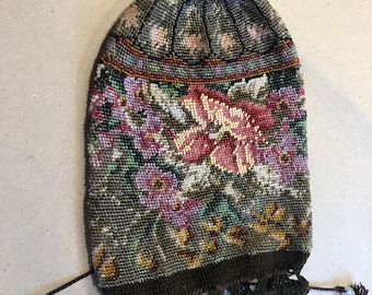 Op de afbeelding: Een vintage kralen trekkoordtas met een bloemenontwerp. De tas heeft een donkergroene achtergrond met roze, paarse en witte bloemen. De bovenkant heeft een kralen lus en de onderkant een donkergroene gehaakte rand. Zwarte trekkoorden zijn zichtbaar.