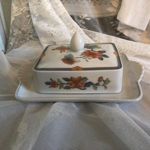 Può includere: Un burriera rettangolare in porcellana bianca con coperchio, decorata con motivi floreali arancioni, verdi e blu. Poggia su un vassoio abbinato con bordo smerlato. Il coperchio ha una piccola maniglia bianca a forma di goccia.