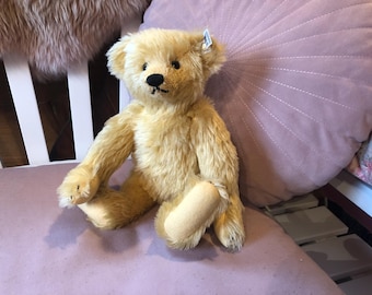 May include: A golden-yellow teddy bear with a black nose and small, dark eyes. The plush toy is sitting on a mauve surface, with a matching pillow in the background. The bear has a tag on its ear.