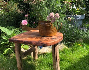 Könnte beinhalten: Ein rustikaler Holztisch mit quadratischer Tischplatte und vier Beinen aus Baumstämmen. Auf dem Tisch steht ein brauner Keramikkrug mit rosa Blüten. Der Tisch befindet sich in einem Garten mit grünem Gras und Blättern.