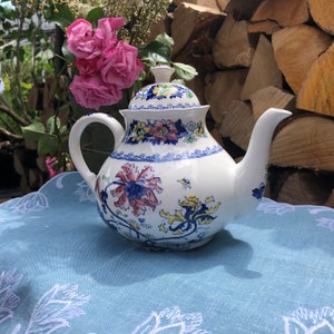 Può includere: Una teiera bianca con un motivo floreale blu e giallo. La teiera ha un coperchio e un manico. La teiera è appoggiata su una tovaglia blu.