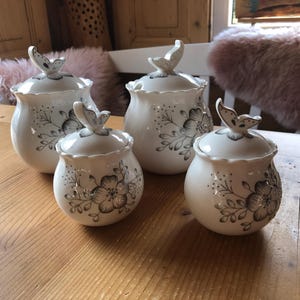 Könnte beinhalten: Vier weiße Keramikbehälter mit schwarzen Blumendesigns und Schmetterlingsakzenten. Die Behälter haben Deckel und sind auf einer Holzoberfläche angeordnet.
