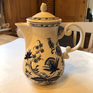 Op de afbeelding: Crèmekleurige keramische koffiepot met deksel, versierd met blauwe bloempatronen. De pot heeft een gebogen handvat en een schenktuit. Het deksel is bekroond met een kleine, knopvormige handgreep. Klassiek ontwerp.