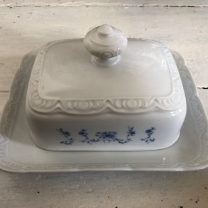 Op de afbeelding: Witte porseleinen botervloot met deksel en bijpassende schaal. De schaal heeft een vierkante vorm met een decoratieve geschulpte rand en een blauw bloemmotief. Het deksel heeft een ronde knop.