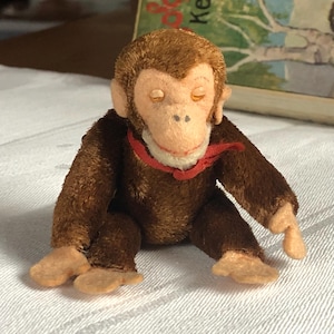 Op de afbeelding: Een vintage pluchen aap met bruine vacht, een lichtgekleurd gezicht en een rode strik om zijn nek. De aap zit met gesloten ogen en zijn armen en benen zijn uitgestrekt. Het speelgoed staat op een wit oppervlak.