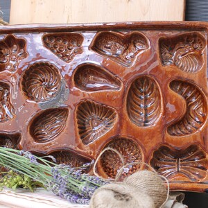 Op de afbeelding: Een bruine keramische bakvorm met 12 individuele vormen. De vormen zijn gevormd als verschillende ontwerpen, waaronder bloemen, bladeren en geometrische vormen. De vorm heeft twee handvatten aan de zijkanten.