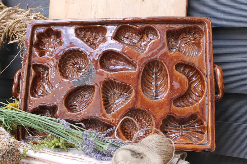 Op de afbeelding: Een bruine keramische bakvorm met 12 individuele vormen. De vormen zijn gevormd als verschillende ontwerpen, waaronder bloemen, bladeren en geometrische vormen. De vorm heeft twee handvatten aan de zijkanten.
