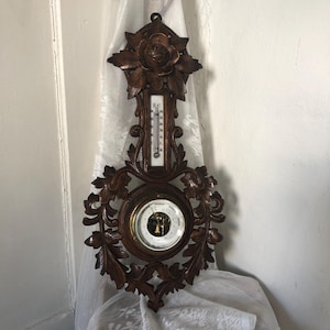May include: A vintage wooden weather station with a thermometer and barometer. The dark brown frame features ornate floral carvings. The thermometer has a white face with black markings. The barometer has a gold-colored face. The weather station is hanging on a wall.