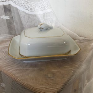 Può includere: Un burriera in porcellana bianca con base rettangolare e coperchio abbinato. Il coperchio presenta una piccola maniglia scolpita ed è decorato con un sottile bordo dorato lungo i bordi. Anche la base ha un bordo dorato, creando un aspetto coeso.