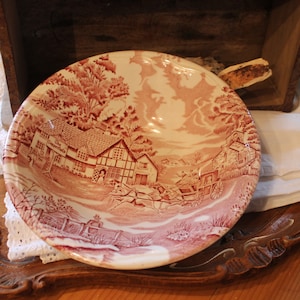 Peut inclure: Un bol en céramique vintage rouge et blanc avec une scène de campagne représentant une maison, des arbres et une calèche tirée par des chevaux. Le bol est sur un plateau en bois avec une dentelle blanche.