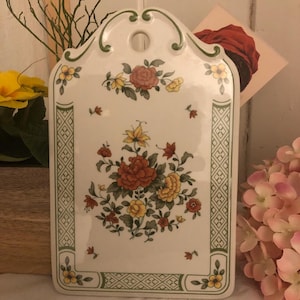 Pode incluir: Uma tábua de cortar de cerâmica branca com uma borda verde e um design floral. O design apresenta flores vermelhas, amarelas e verdes.