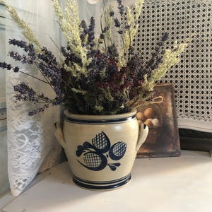 Puede incluir: Un jarrón de cerámica color crema con diseños florales azules, lleno de lavanda y trigo secos. El jarrón tiene dos asas y un borde azul. Una pintura de naturaleza muerta de estilo vintage está en el fondo.