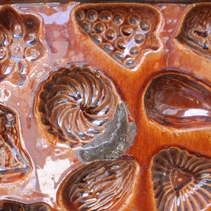 Op de afbeelding: Een vintage bruine keramische vorm met meerdere holtes om verschillende vormen te bakken, waaronder een bloem, een werveling, een tros druiven, een aubergine en een hart.