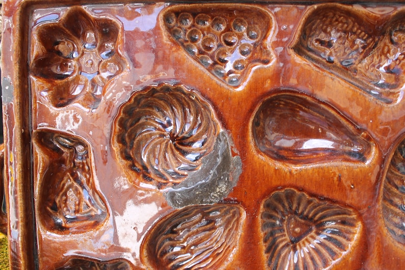 Op de afbeelding: Een vintage bruine keramische vorm met meerdere holtes om verschillende vormen te bakken, waaronder een bloem, een werveling, een tros druiven, een aubergine en een hart.