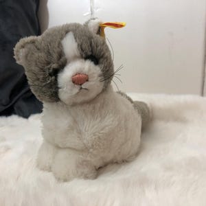 Könnte beinhalten: Ein graues und weißes Plüschkatzenspielzeug mit weißem Gesicht und rosa Nase. Die Katze hat ein kleines gelbes Etikett am Ohr.