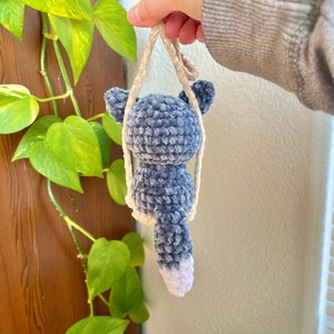 May include: A blue and white crocheted plush toy shaped like a cat with a long tail. The toy is hanging from a white crocheted string.