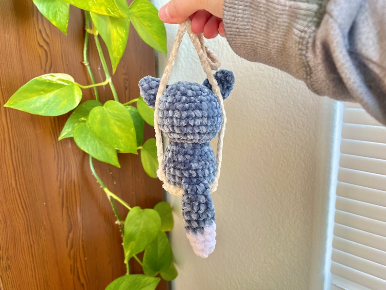 May include: A blue and white crocheted plush toy shaped like a cat with a long tail. The toy is hanging from a white crocheted string.