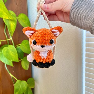 May include: A small orange and white crocheted fox keychain with black eyes and a white nose. The fox is hanging from a white cord.