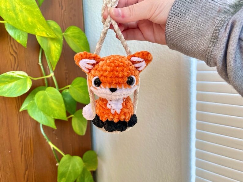 May include: A small orange and white crocheted fox keychain with black eyes and a white nose. The fox is hanging from a white cord.