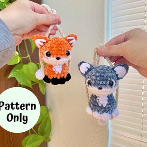 以下が含まれることがあります： 2つのアミグルミのおもちゃを作るための編み図です。キツネはオレンジと白、犬はグレーと白です。両方の玩具は紐で吊り下げられています。