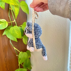 May include: A blue and white crocheted plush toy shaped like a cat hanging from a beige cord. The toy has a long tail and white accents on its face and paws.