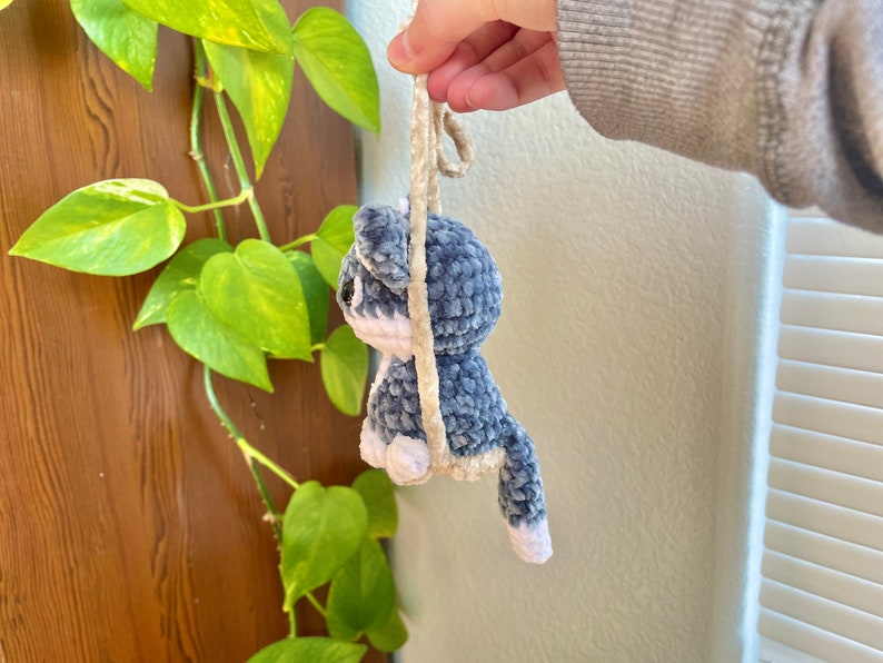 May include: A blue and white crocheted plush toy shaped like a cat hanging from a beige cord. The toy has a long tail and white accents on its face and paws.