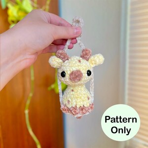 May include: A handmade crochet giraffe in yellow and brown, held by a hand. The giraffe has large black eyes and small brown horns. The text "Pattern Only" is in a green circle.