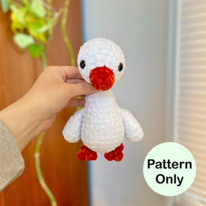 May include: A handmade white crochet goose with a red beak and feet. The goose has black eyes and small wings. The text "Pattern Only" is in a green circle in the lower right corner. The goose is a soft, plush toy.