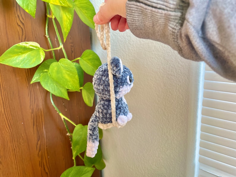 May include: A crocheted plush toy in the shape of a cat, with a blue and white color scheme. The cat is hanging from a white string.