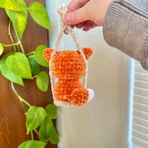 May include: A small orange crocheted fox toy hanging from a white string. The fox is sitting on a swing and has a white tail.