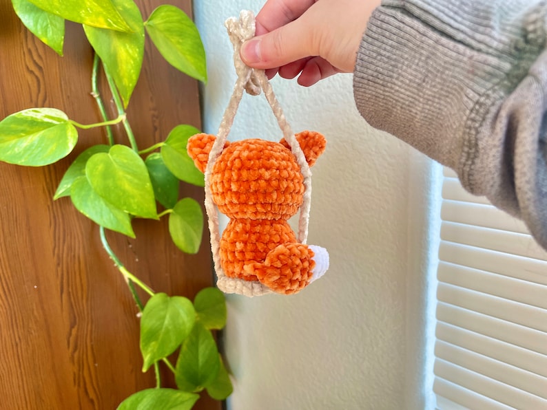 May include: A small orange crocheted fox toy hanging from a white string. The fox is sitting on a swing and has a white tail.