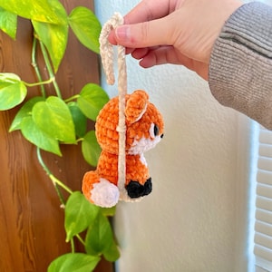 May include: A crocheted orange fox with white accents hanging from a white string. The fox has black eyes and a black nose.