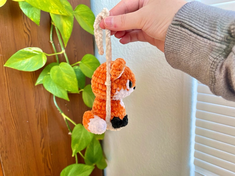 May include: A crocheted orange fox with white accents hanging from a white string. The fox has black eyes and a black nose.