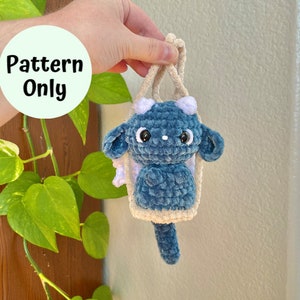 Puede incluir: Un juguete de peluche azul y blanco tejido a crochet con forma de dragón con ojos grandes. El dragón está colgado de una bolsa de crochet de color crema.