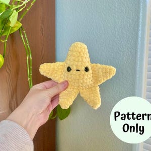 Op de afbeelding: Een gehaakt geel knuffelsterretje met zwarte ogen. Het speelgoed wordt vastgehouden door een hand voor een blauwe muur. De tekst "Pattern Only" is in zwart op een witte cirkel geschreven.