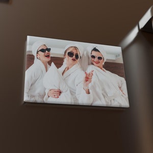 May include: Three women wearing white bathrobes and sunglasses are smiling and posing for a photo. The photo is printed on a white square magnet with rounded corners.