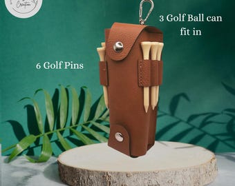 May include: A brown leather golf ball and tee holder with a silver clasp. The holder has slots for six golf pins and can fit three golf balls. The holder is on a wooden stand against a green background.
