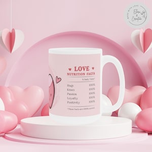 May include: White ceramic mug with a pink design featuring the text "LOVE NUTRITION FACTS" and a list of love-related items with a percentage value of 100%. The mug has a pink heart design and a pink handle.