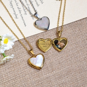 Puede incluir: Dos collares de oro con colgantes en forma de corazón. Un colgante tiene un interior de nácar blanco, y el otro colgante tiene una foto de una persona sosteniendo un perro. El colgante con la foto tiene la inscripción "Forever in my heart".