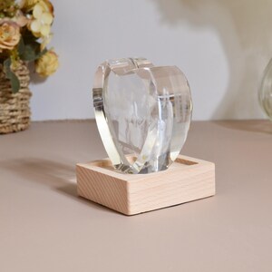 May include: A clear glass heart-shaped ornament with a photo inside, sitting on a wooden base. The photo is of a group of people and the text "Te amo" is visible.