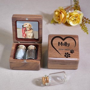 Op de afbeelding: Een houten herdenkingsdoos met een foto van een golden retriever erin. De doos is open en onthult twee kleine glazen potjes. Een tweede doos is gesloten, met de naam "Molly", data en pootafdrukken gegraveerd op de voorkant. Een kleine glazen fles met een kurk ligt ervoor.