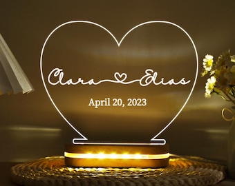 May include: A heart-shaped LED night light with the names "Clara & Elias" and the date "April 20, 2023" etched in white. The light sits on a wooden base and is placed on a woven mat. The light emits a warm glow.