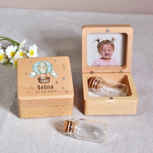 Puede incluir: Dos cajas de recuerdos de madera. Una caja cerrada presenta un diseño de globo aerostático con un oso de peluche, el nombre "Selina" y la fecha "25.03.2024". La otra caja está abierta, mostrando una foto y una pequeña botella de vidrio con un corcho.