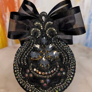 May include: A black fabric brooch with a large, ornate, black rhinestone design. The brooch is accented with a black satin bow.