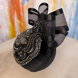 May include: A black velvet ornament with a black satin bow and iridescent rhinestones. The ornament is decorated with a floral design and is perfect for adding a touch of elegance to any holiday decor.