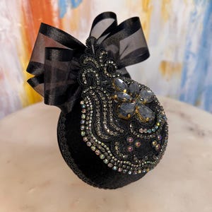 May include: Black velvet Christmas ornament with a black satin ribbon and a large iridescent jewel in the center. The ornament is decorated with black beads and sequins.