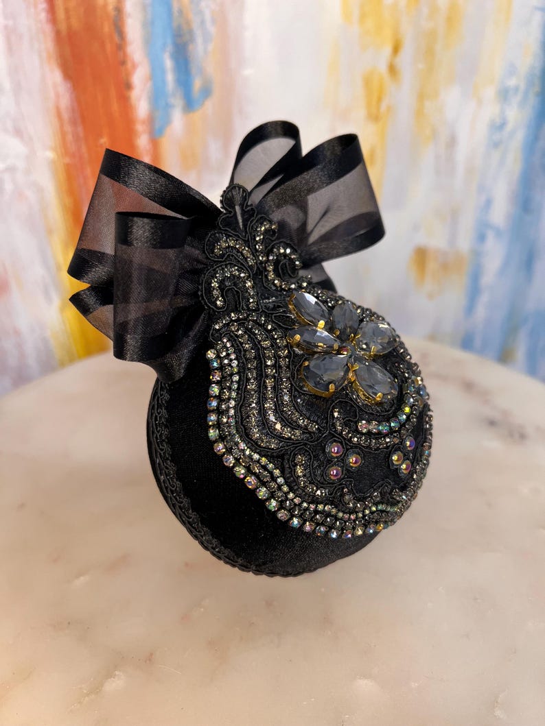 May include: Black velvet Christmas ornament with a black satin ribbon and a large iridescent jewel in the center. The ornament is decorated with black beads and sequins.