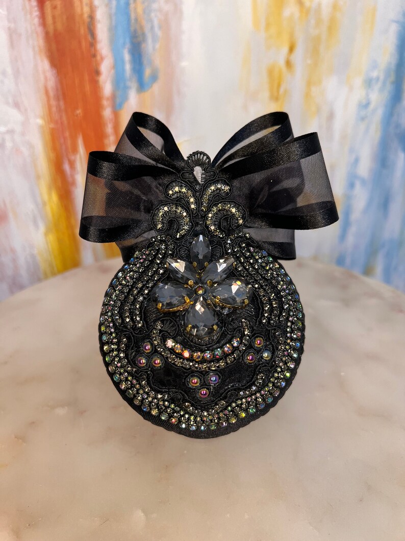 May include: A black fabric brooch with a large, ornate, black rhinestone design. The brooch is accented with a black satin bow.